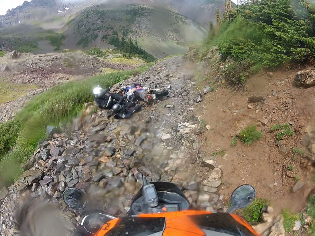 Una moto tumbada y dos personas en el suelo, sobre un camino de piedras