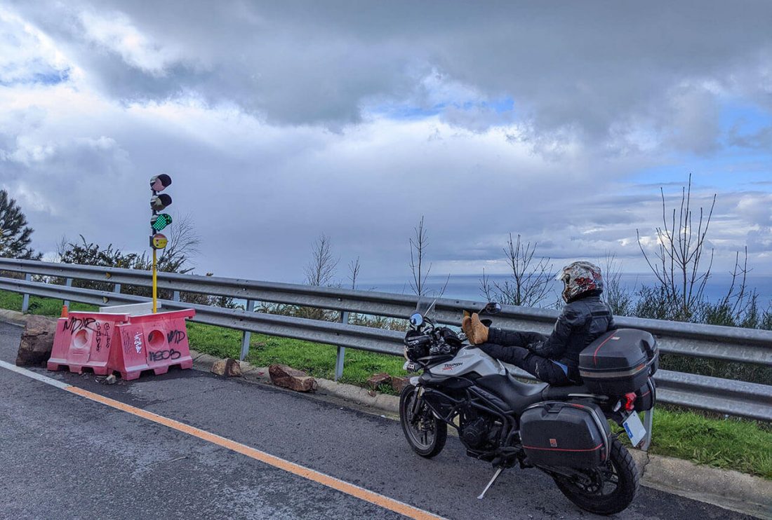 Moto parada en un semáforo de obra en carretera de montaña con mar y cielo nuboso